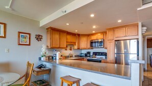 A kitchen with wooden cabinets, a stainless steel refrigerator, a microwave, and a coffee maker. There is a small island with stools and a chair.