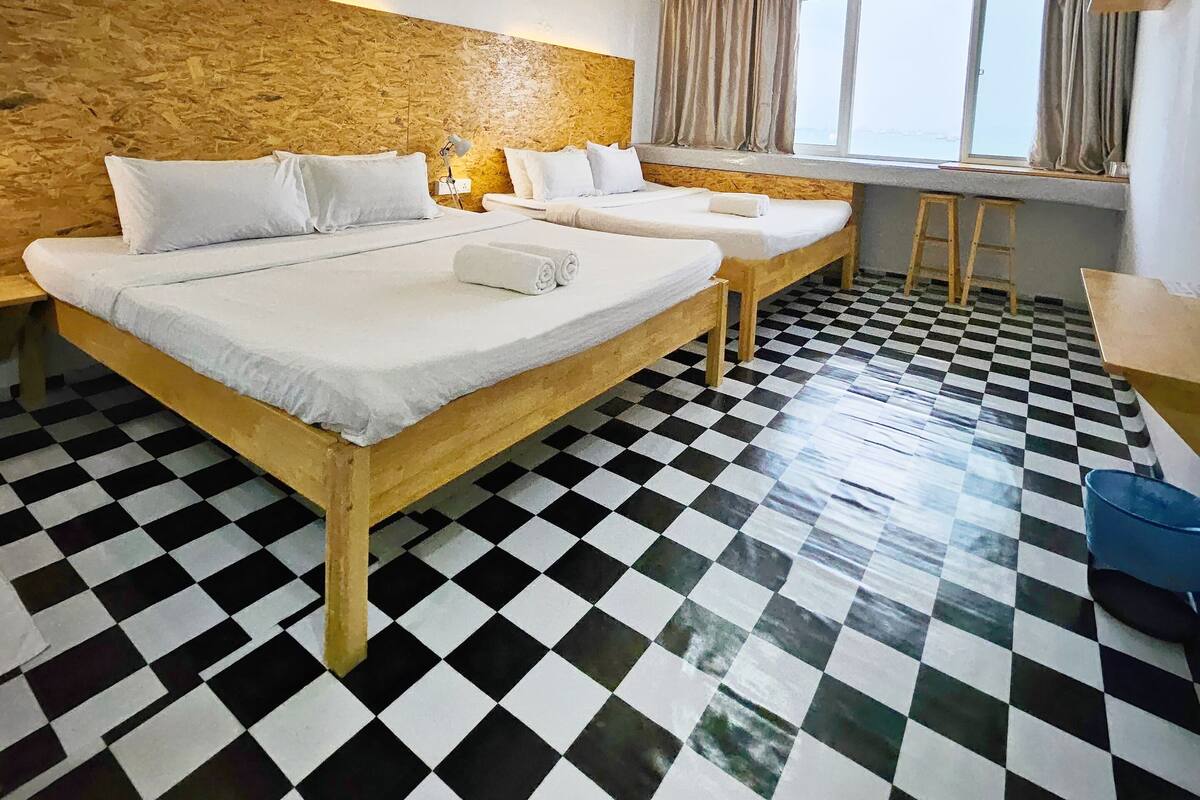 A hotel room with two beds, checkered floor, wooden headboard, large window with curtains, and a small desk.
