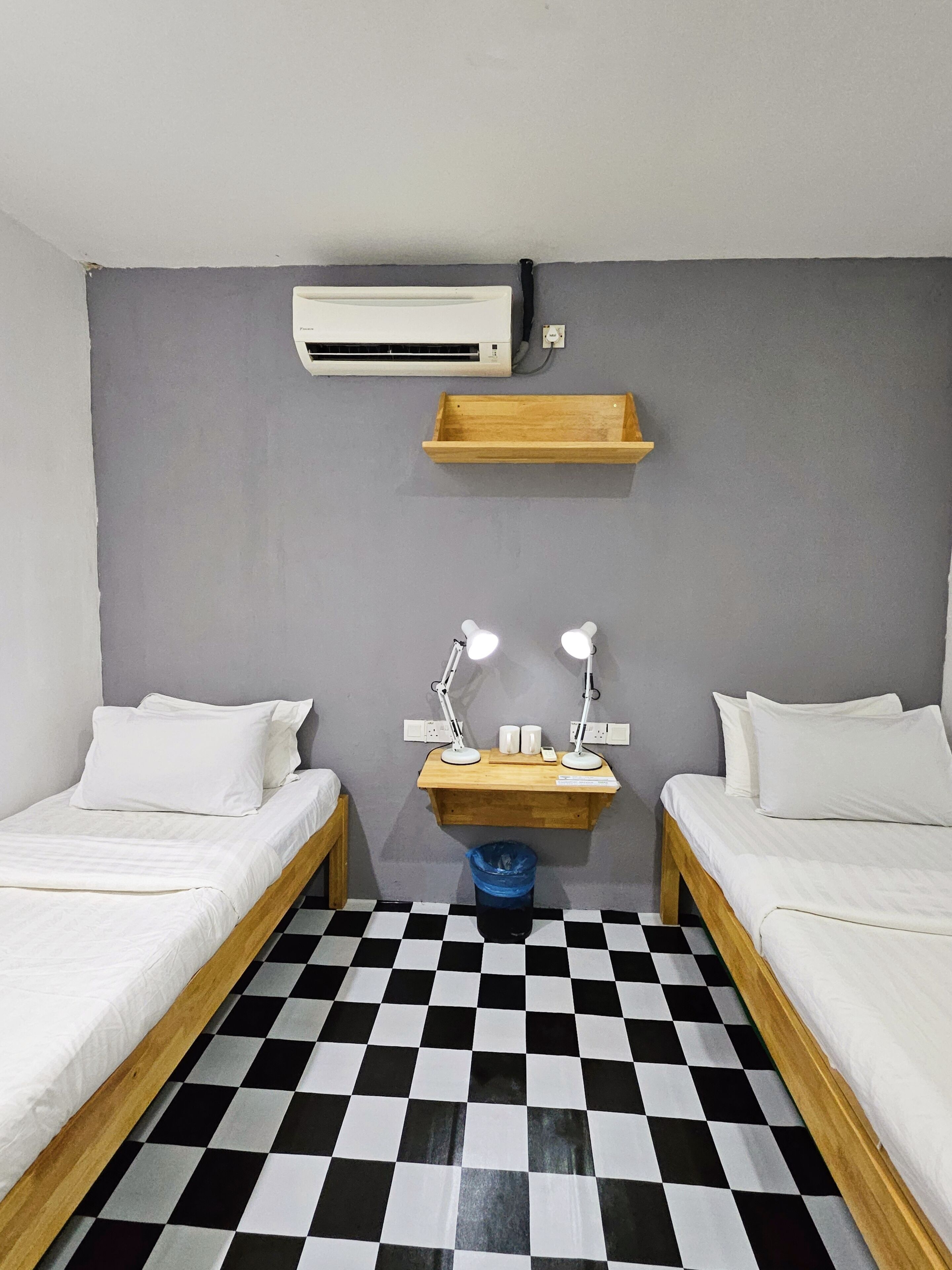 A room with two single beds, a small wooden shelf, a wall-mounted air conditioner, and a checkered floor.