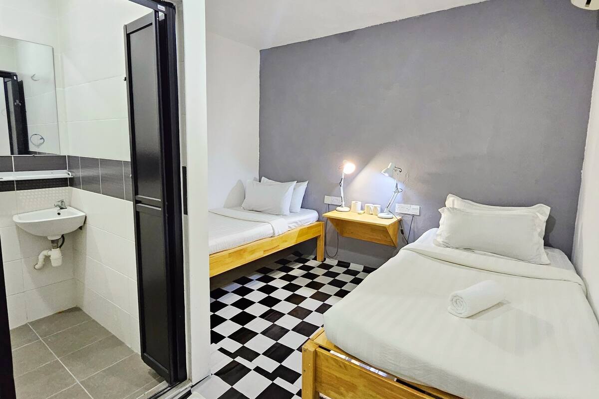 A hotel room with a checkered floor, a bed, a bedside table with a lamp, a small sink, and a mirror.