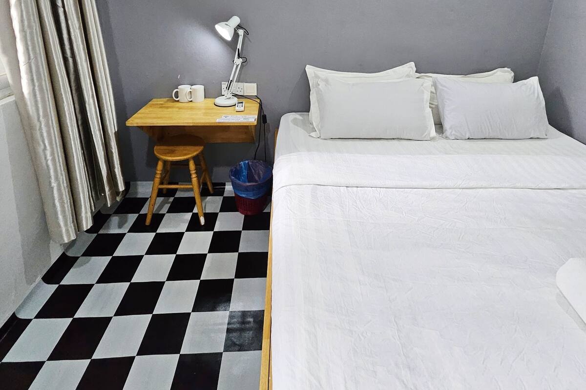 A hotel room with a checkered floor, a bed with white linens, a desk with a lamp, and an air conditioner mounted on the wall.
