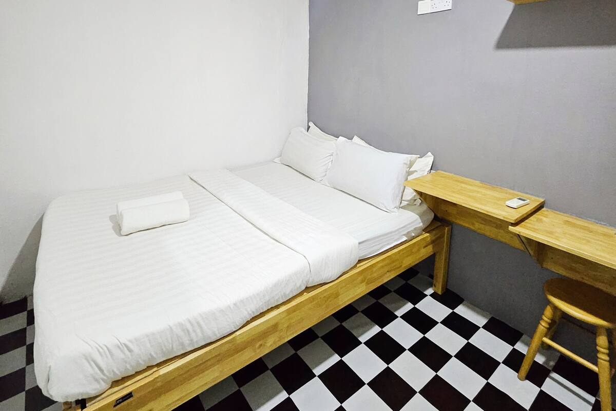 A single bed with a wooden frame, a small wooden desk, a chair, a wall-mounted air conditioner, and a checkered floor.