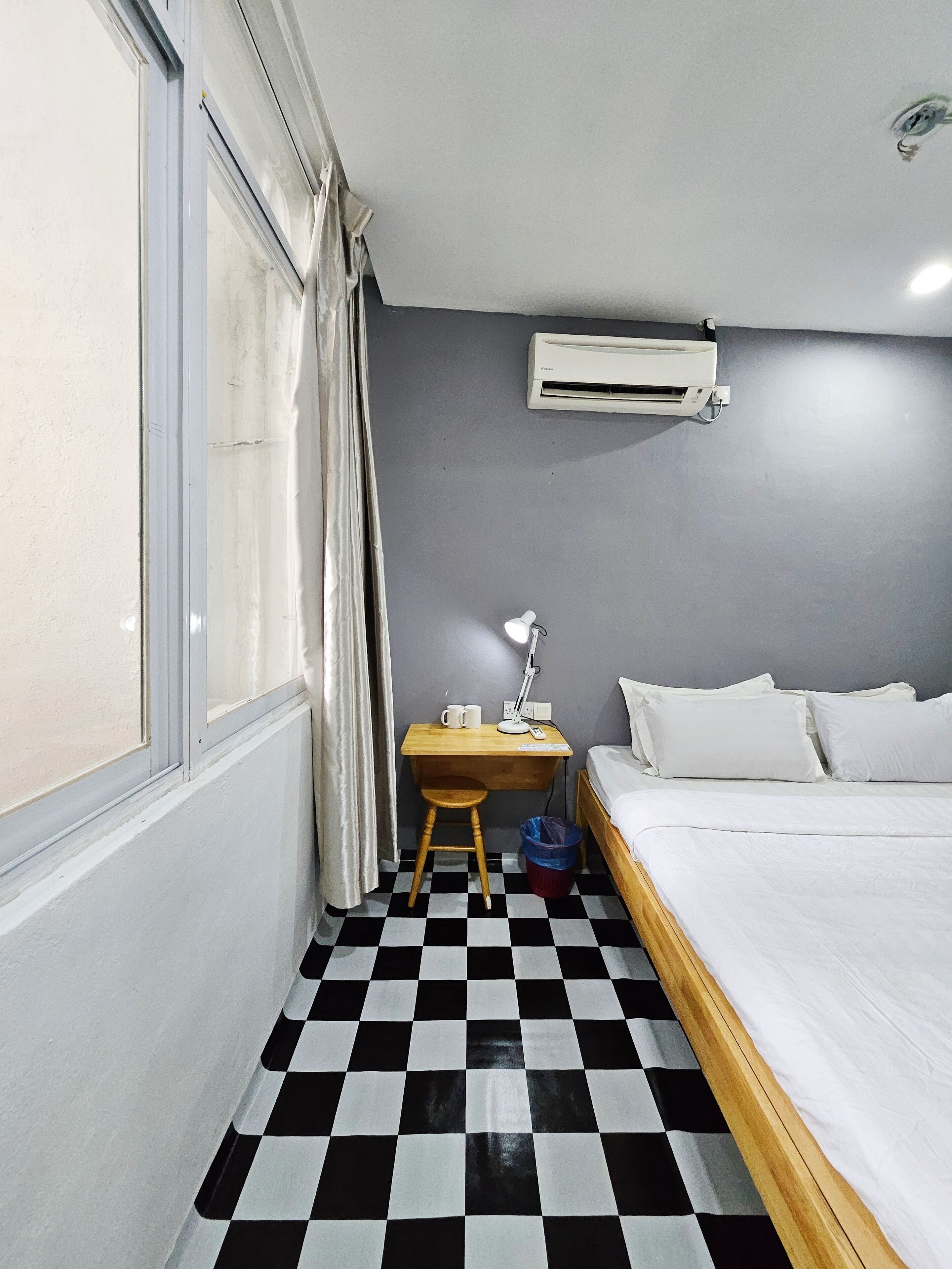 A small, neatly arranged bedroom with a checkered floor, a bed, a desk with a lamp, a chair, and an air conditioning unit.