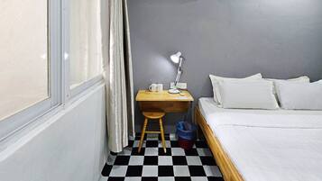 A small, neatly arranged bedroom with a checkered floor, a bed, a desk with a lamp, a chair, and an air conditioning unit.