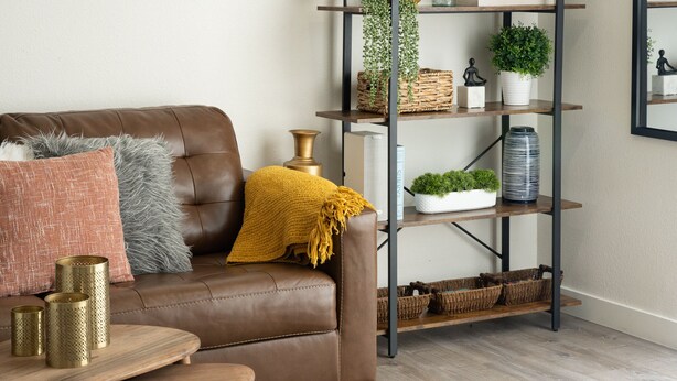 A brown leather sofa with a mustard throw blanket, a small wooden side table with three gold-colored cylindrical objects, and a metal shelving unit with baskets and plants.