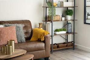 A brown leather sofa with a mustard throw blanket, a small wooden side table with three gold-colored cylindrical objects, and a metal shelving unit with baskets and plants. - Plush King Bed, Full Kitchen, Fast WiFi, Workspace, Parking (Denver)