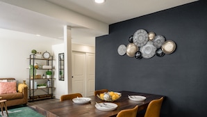A dining area with a dark wall, a wooden table, and yellow chairs. There is a fruit basket on the table and a bookshelf with various items in the background.