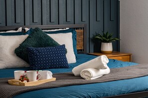 Un lit bien fait, recouvert d’une couette bleue, d’un plaid tricoté gris et d’une serviette blanche roulée. Sur un plateau, on aperçoit deux tasses blanches, un petit objet décoratif rouge et blanc, et une plante en pot posée sur une table de chevet en bois, à côté du lit.
