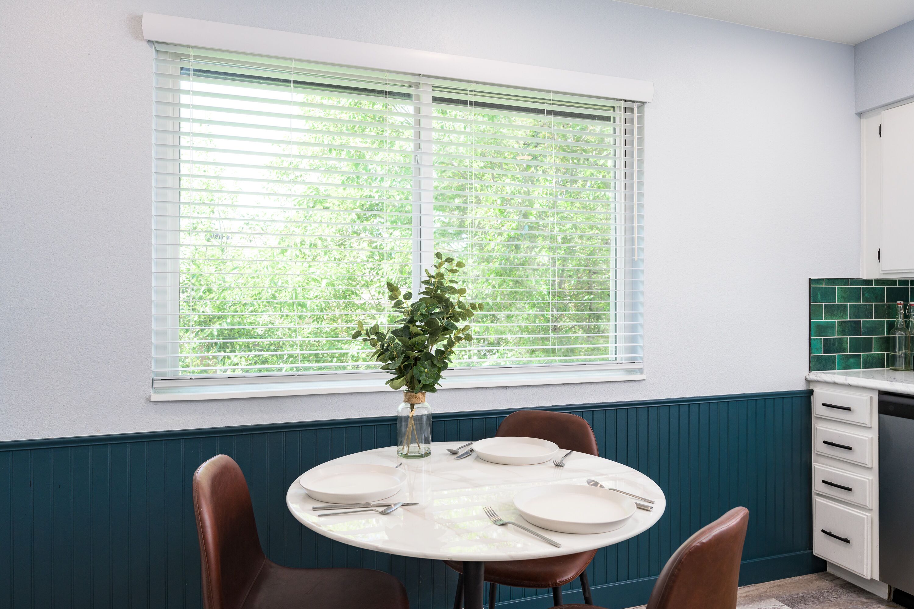 A dining area with a round table set for four, a vase with a plant, and a window with blinds.