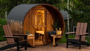 Eine Holzsauna mit gewölbtem Dach, zwei Adirondack-Stühlen und einem Tisch mit Eimer und Handtuch.