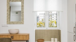 A room with a wooden table, a white bowl, a mirror, a window with a view of buildings, and a shelf with two containers. - Casa Buganvília (Funchal)