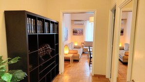 A hallway with a black bookshelf, a potted plant, and a wooden floor leading to a living area with a sofa, a table, and a lamp. - Comfortable apartment in Syntagma square (Athina)