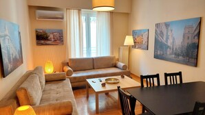 A cozy living room with a sofa, a coffee table, and a dining table with chairs. There are paintings on the walls and an air conditioning unit above the sofa. - Comfortable apartment in Syntagma square (Athina)