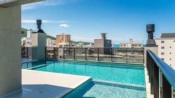 A rooftop swimming pool with clear blue water, surrounded by modern buildings and a view of a hilly landscape.