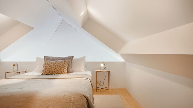 A bedroom with a sloped ceiling, a bed with a beige bedspread, two bedside tables with lamps, and a view of the sky through the ceiling. - Atelier by the Sea (Porto)
