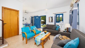 A living room with a wooden door, blue armchairs, a grey sofa, a small table with a placemat, and a fan. - Villa Rotiana - Three Bedroom Villa, Sleeps 6 (Poli Crysochous)