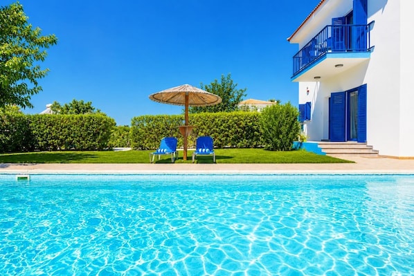 Piscina cristalina con sillas azules y una sombrilla, rodeada de follaje y un edificio blanco con detalles azules.