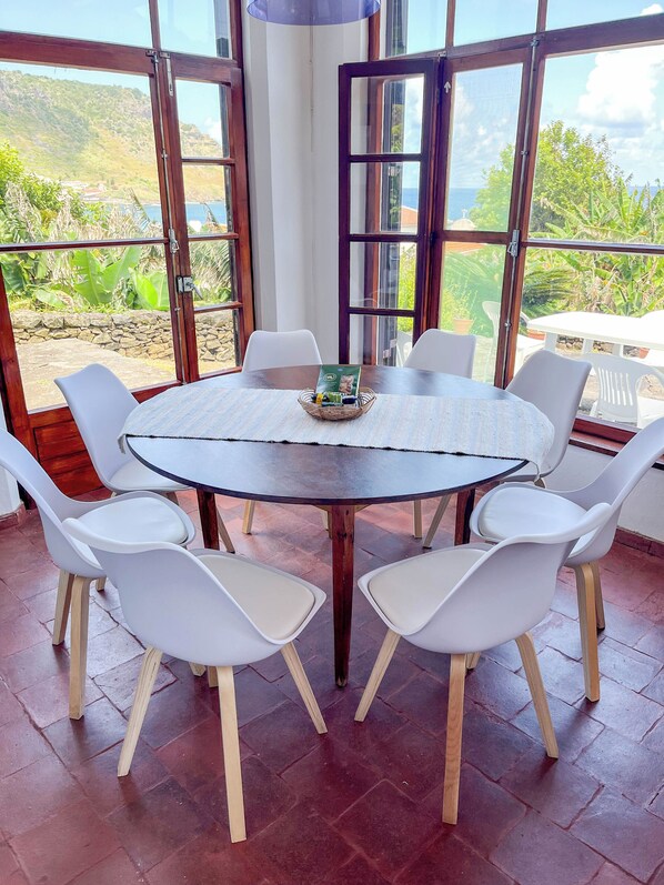 A round table with white chairs, a basket with items, and a view of greenery through large windows. - Casa dos Magalhães (Vila do Porto)