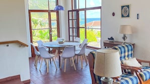 A spacious room with a wooden ceiling, large windows offering a view of greenery and the sea, a dining area with a table and chairs, and a sofa with a lamp. - Casa dos Magalhães (Vila do Porto)