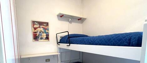 A small, single-bedded room with a white bunk bed, a small shelf, and a wall-mounted picture.