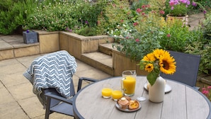 A round wooden table with a blue and white patterned throw, two glasses of orange juice, a pitcher, a plate with muffins, and a vase with sunflowers.