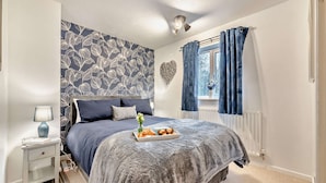 A bedroom with a patterned wallpaper, a bed with a grey blanket, a bedside table with a lamp, and a window with blue curtains.