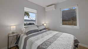 A bedroom with a bed, two bedside tables with lamps, an air conditioning unit, and two windows offering views of the outdoors. - Lake Front New Remodel w/ Hot Tub- Coeur d'Alene (Coeur d'Alene)