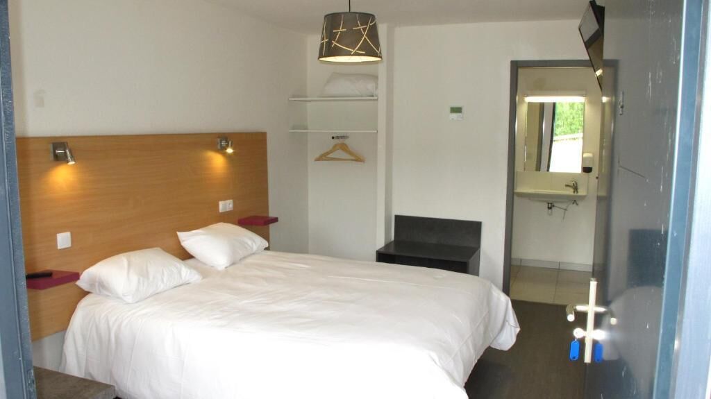 Une chambre d’hôtel comprenant un lit, des tables de chevet, un luminaire suspendu, un banc, un miroir et une porte donnant accès à une salle de bain.