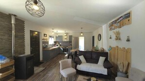 Un salon moderne avec un canapé, des fauteuils et une table basse. On y trouve une cheminée, une cuisine à l’arrière-plan et un coin repas.