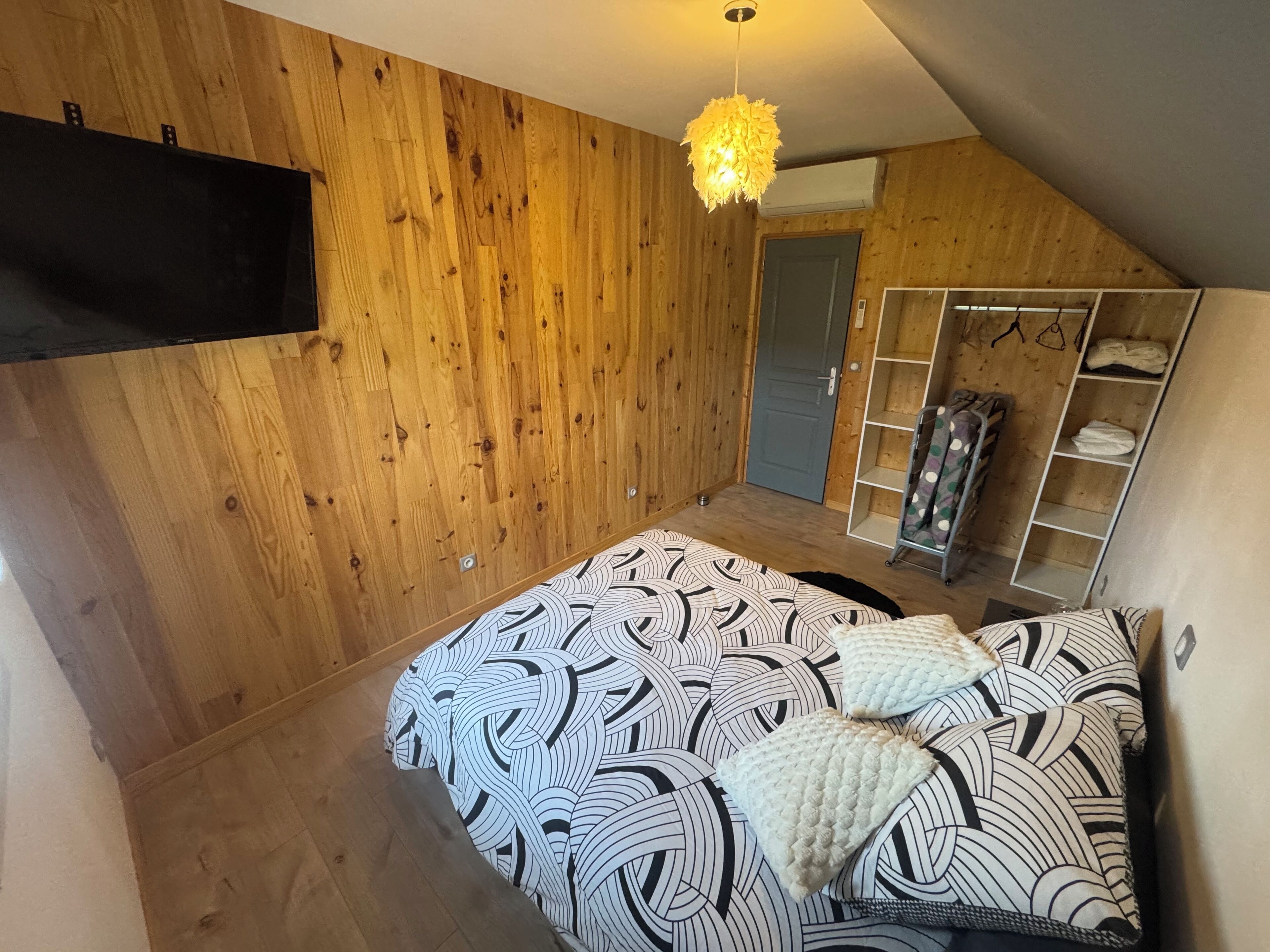 Un dormitorio con techo inclinado, paredes de madera, cama con ropa de cama estampada, lámpara colgante y vestidor con estantes.