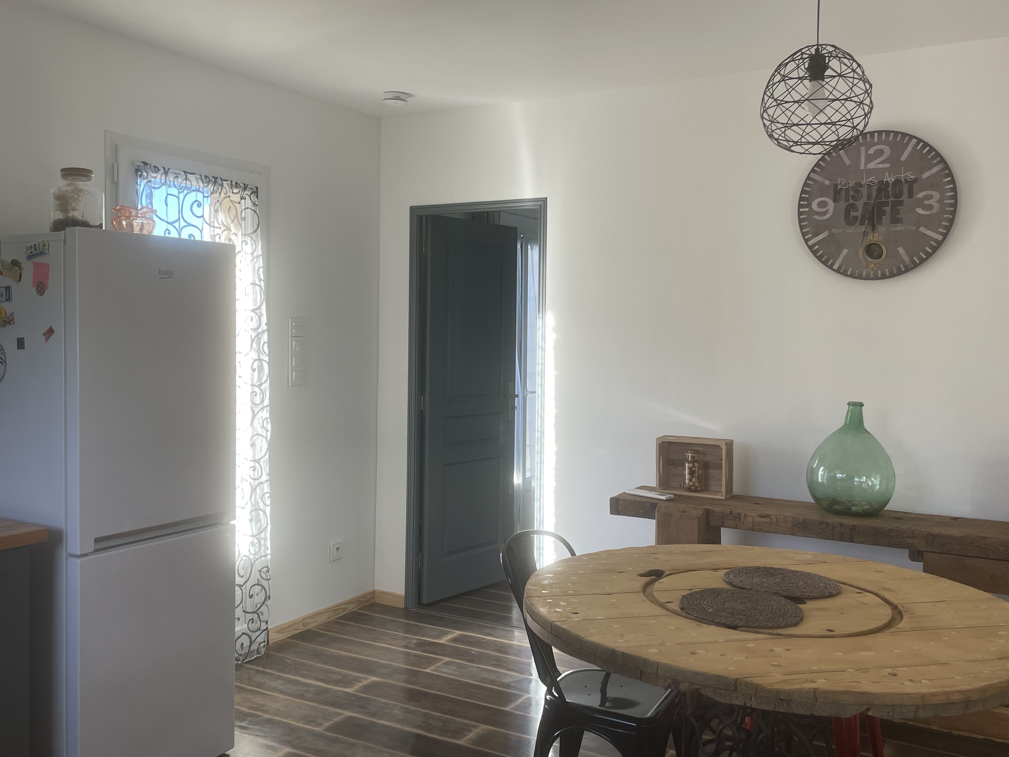 Une cuisine équipée d’un réfrigérateur, d’une table à manger avec des chaises, d’une horloge au mur et d’une porte donnant sur une autre pièce.