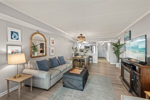 A modern living room with a sofa, ottoman, and coffee table, a dining area with a table and chairs, and a kitchen in the background.