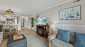 Una sala de estar moderna con un sofá, una mesa de centro y un televisor. Se observa una cocina al fondo con una zona de comedor.