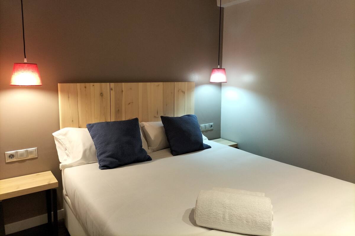 A neatly made bed with a white comforter and two pillows, a wooden headboard, and a bedside table with a lamp.