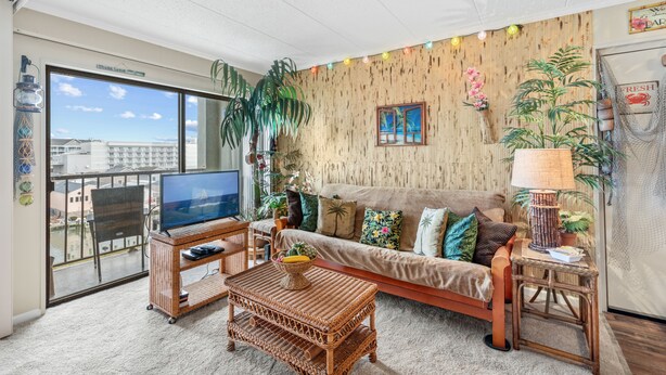 A living room with a large TV, wicker furniture, a sofa with colorful cushions, and a view of buildings through a large window. - Westward Ho 410 by Seaside Vacations & Sales (Ocean City)