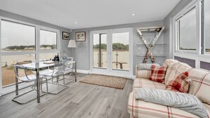A modern living room with a glass dining table, a sofa with plaid cushions, and large windows offering a view of the outdoors. - The Old Pilot Hut, Felixstowe Ferry, Amazing Views (Felixstowe Ferry)