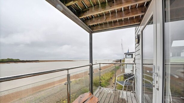 A covered outdoor balcony with a wooden bench, a metal railing, and a view of a river. - The Old Pilot Hut, Felixstowe Ferry, Amazing Views (Felixstowe Ferry)