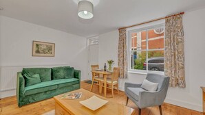 A living room with a green sofa, a grey armchair, a wooden coffee table, and a dining table with chairs. There is a window with curtains and a painting on the wall.