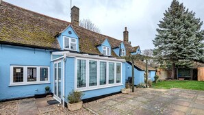 A blue house with a conservatory, a tiled patio, and a large evergreen tree. - Charming Cottage in Barton Mills (Barton Mills)