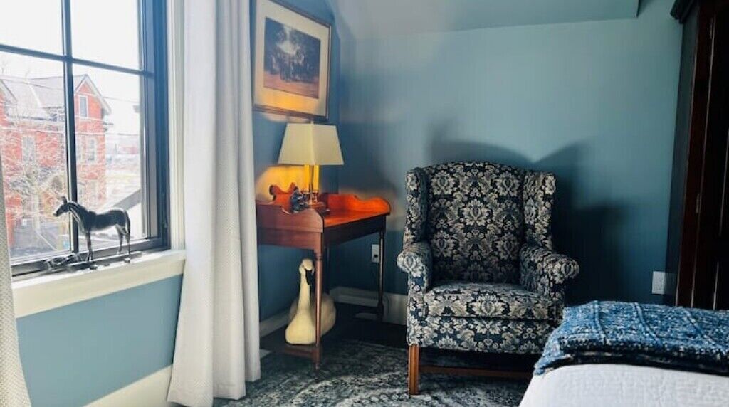 Ein Zimmer mit einem Bett, einem gemusterten Sessel, einem Schreibtisch mit Lampe und einem Fenster mit Blick auf ein Gebäude.