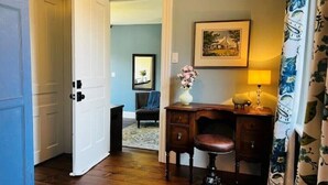 Una habitación con un escritorio de madera, una silla, un jarrón con flores, una lámpara y un cuadro en la pared.