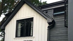Una casa moderna de dos pisos con fachada blanca y gris oscura, puerta principal con marco negro y dos luces exteriores encendidas.