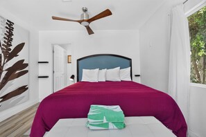 A bedroom with a bed, a ceiling fan, a window with a view of trees, and a door.