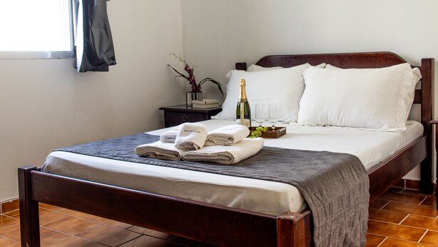 A neatly made bed with white linens, a wooden headboard, and a bedside table with a bottle of champagne, glasses, and a vase with flowers. - Apartamento aconchegante - Caraguatatuba (Caraguatatuba)