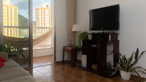 A living room with a large window, a flat-screen TV, a sofa, a small table with a lamp, and a potted plant. - Apartamento aconchegante - Caraguatatuba (Caraguatatuba)