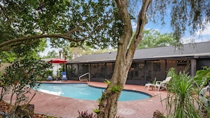 A swimming pool with a brick patio, surrounded by trees and plants, and a building with large windows in the background.