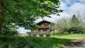 Eine Holzchalet mit einem Balkon, umgeben von üppiger Vegetation und blauem Himmel.