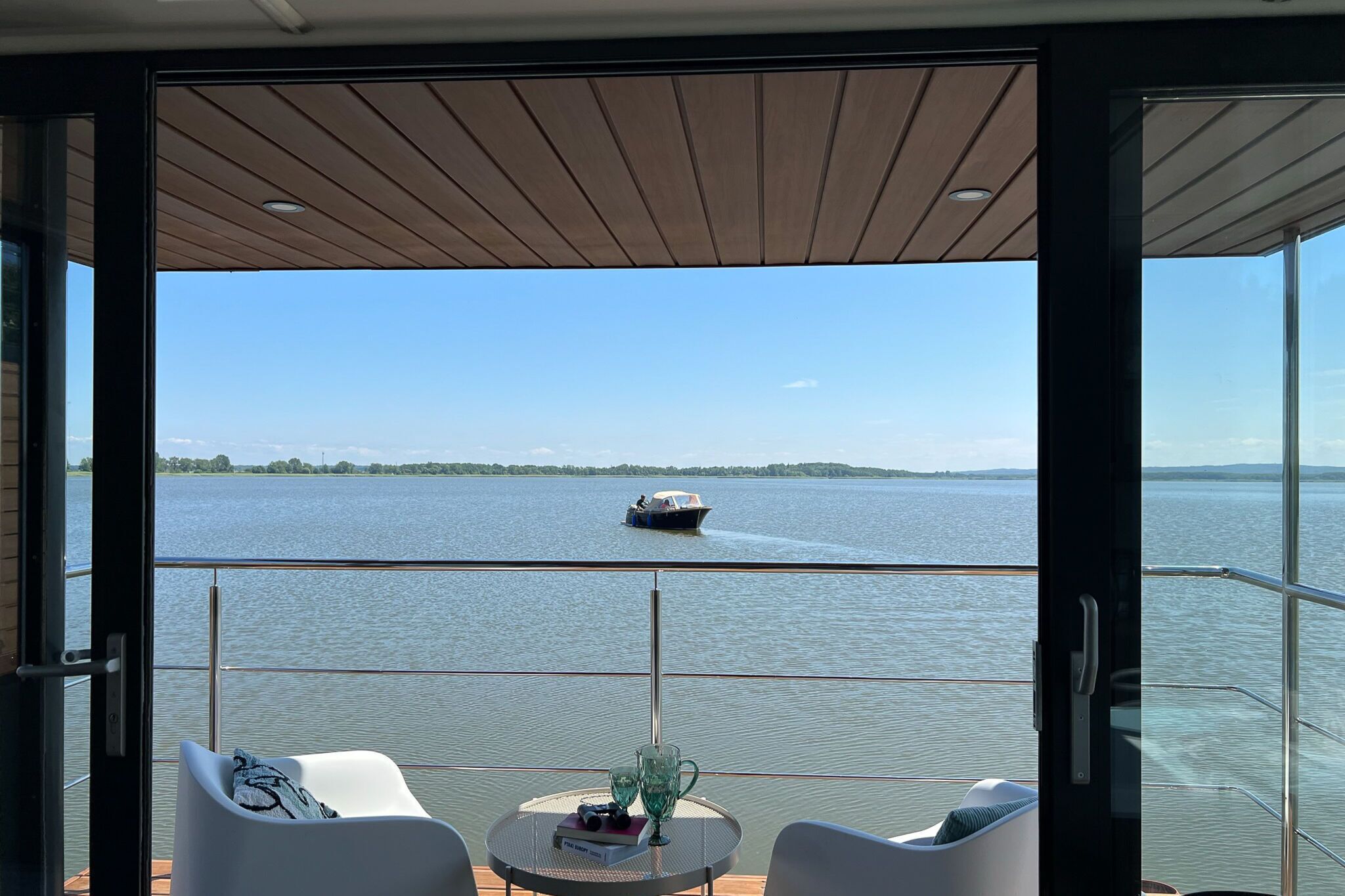 Un balcón con vistas a un lago y un bote. Hay dos sillas y una mesa con un vaso y un libro.