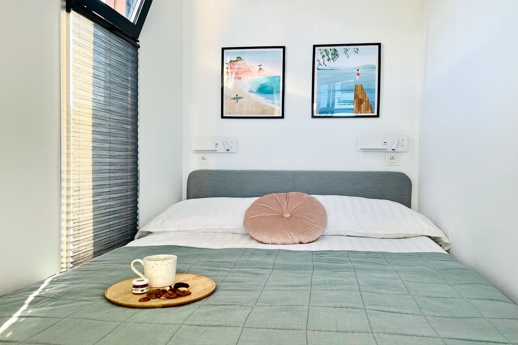 Ein ordentlich bezogenes Bett mit einer grünen Steude, einem rosafarbenen Kopfkissen und einem Tablett mit einer Tasse und Snacks. An der Wand hängen zwei gerahmte Bilder: eines zeigt eine Strandansicht, das andere eine Person, die von einer Klippe springt.