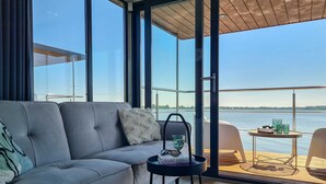 Un salon moderne avec un canapé gris, une petite table ronde sur laquelle se trouvent un livre et des lunettes, et une vue sur une étendue d’eau par de grandes fenêtres.